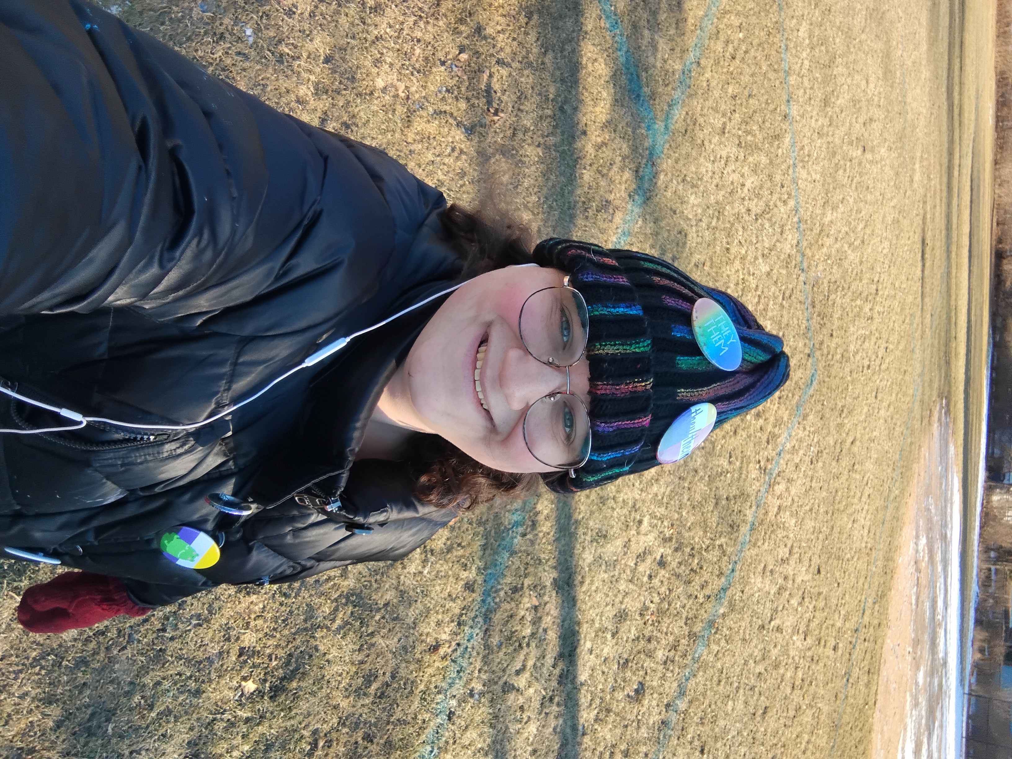 Me, a white enby with brown curly hair and large circular classes. I'm smiling and looking into the camera and wearing a black-rainbow hat with pronoun pins on it and a black shiny coat. I'm standing on a sports field with some snow on the ground in the background.