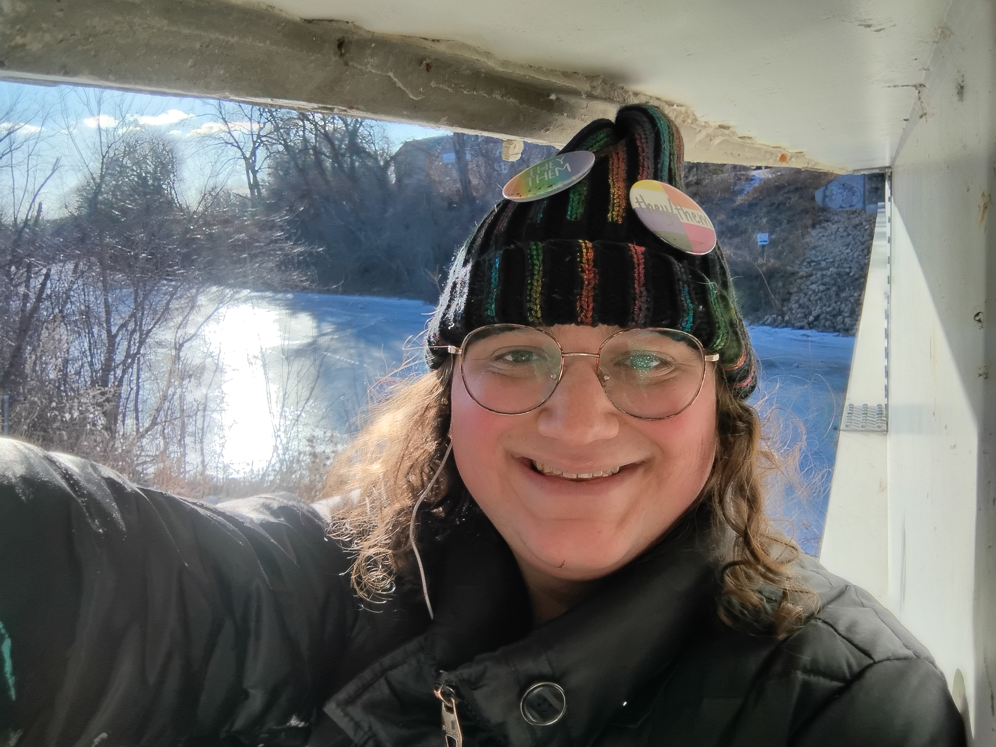 I'm standing on the side support of the bridge, with the side of the bridge on the right, my head almost touching to upper side of the bridge support, and a wide landscape behind me, emphasizing the blue curve of the river with the sun glinting off it.