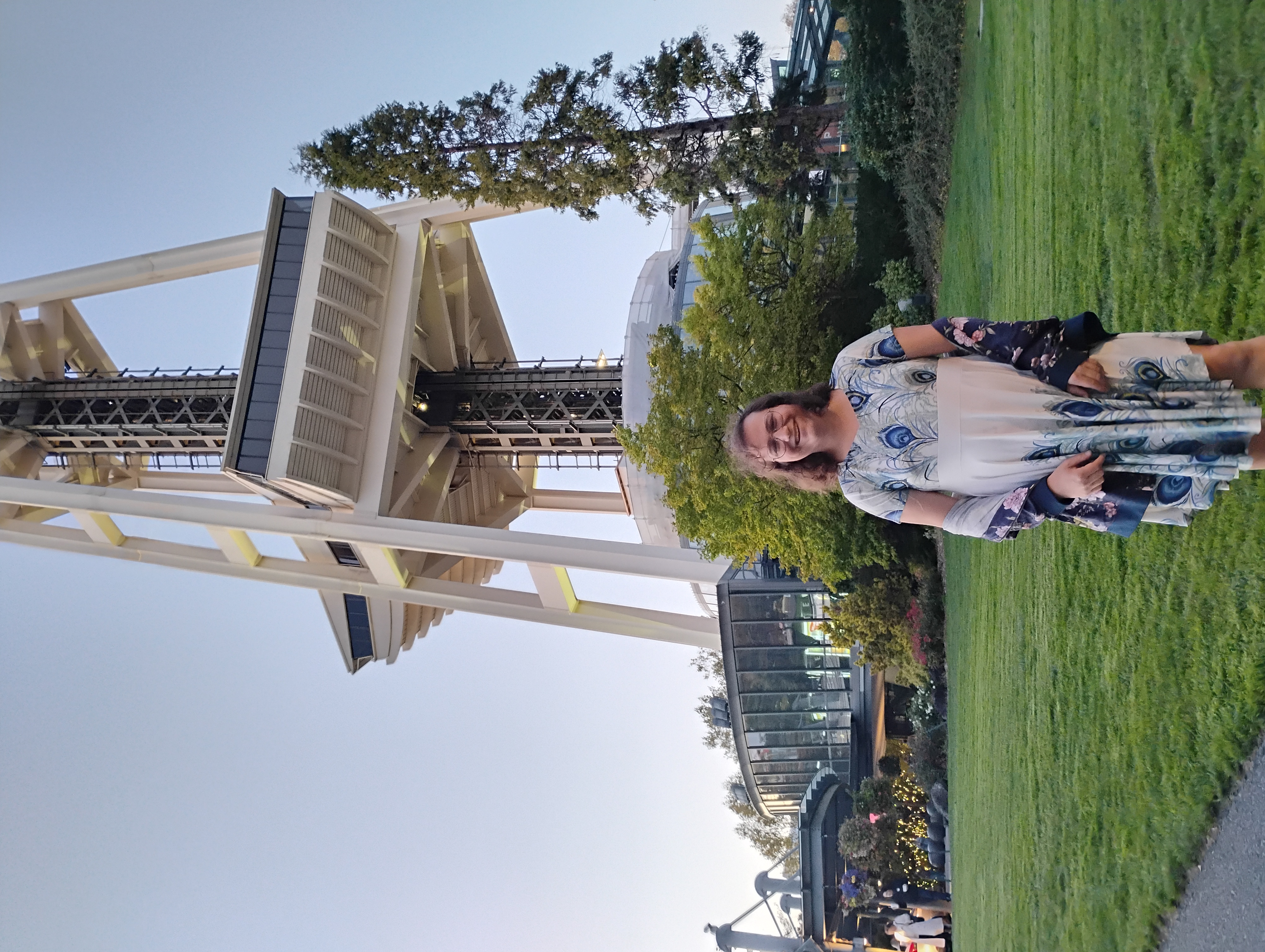 Me, an white enby with dark-brown hair wearing wide-lensed classes, smiling and wearing a peacock dress on a lawn in front of the base of the Space Needle