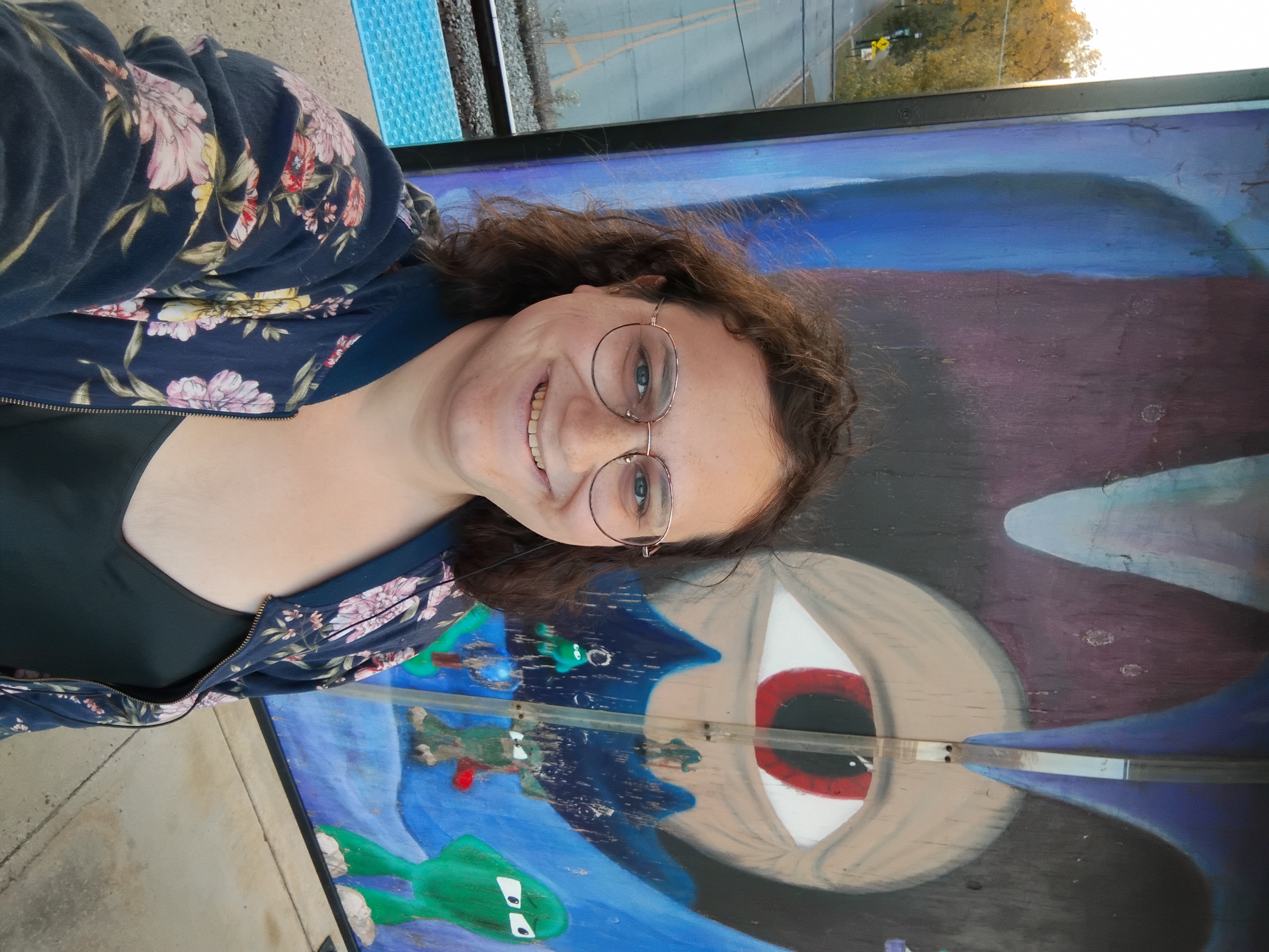 Me wearing a flower-pattern sweater over a black tank-top in front of a large public artwork, with a large eye looking out of a mouth-cave