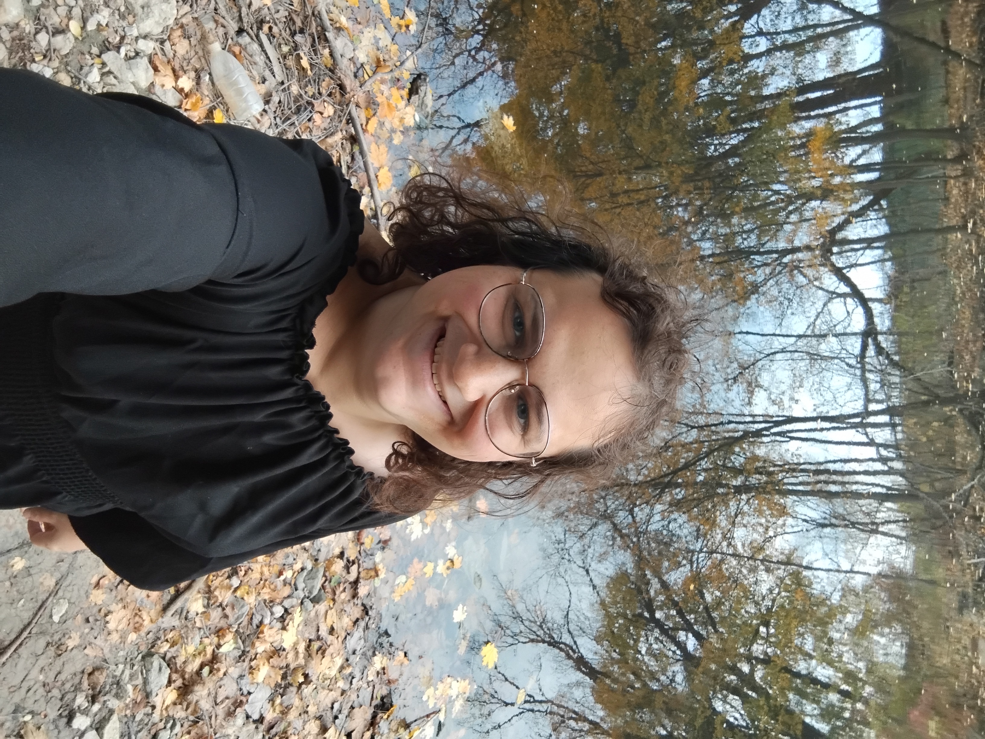 Me, a white ency with brown curly hair, wearing a flowy black top with a wide neckline, in front of a river with yellow fallen leaves. Photo taken from above.