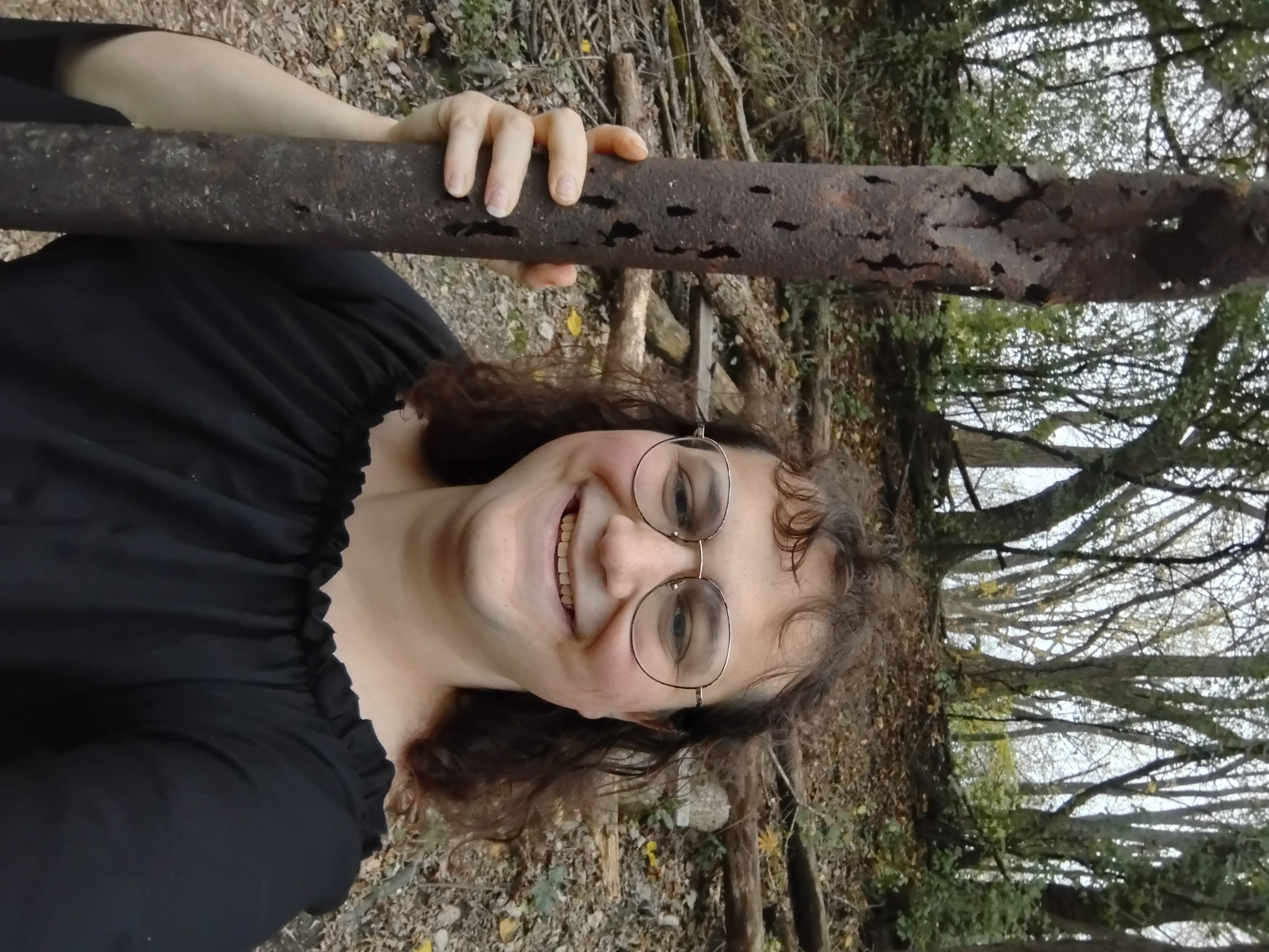 Me holding a tall rusted-out pipe, with corrosion holes and light peeking through it, in front of a haphazard pile of logs