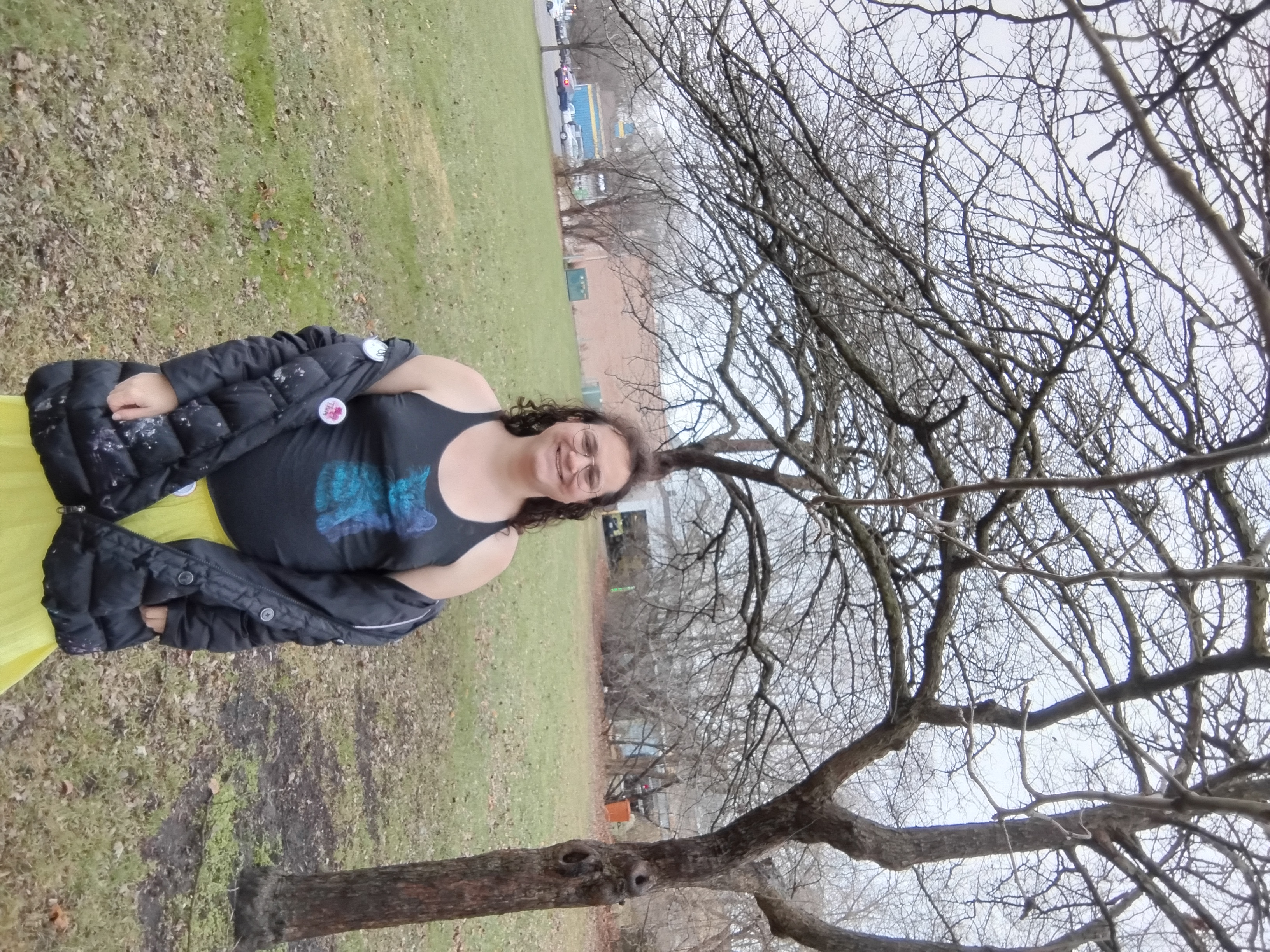 Me, a white enby with brown, curly, shoulder length hair, smiling. I'm wearing a black tanktop with a blue-purple cat on it, a black pufy coat down around my elbows, and a bright yellow skirt. I'm standing in a field in front of a bare-branched tree with a flat grey sky.