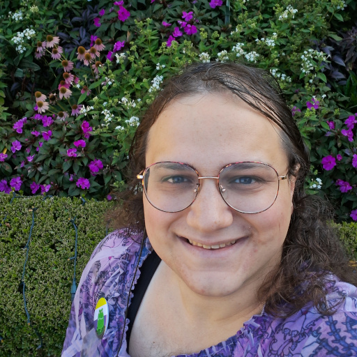 Isaac Grosof's portrait - A nonbinary person with their head and shoulders visible, wearing a purple top in front of a flowerbed.